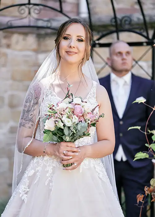 Onelovephoto Eine Braut in einem weißen Spitzenhochzeitskleid hält einen Blumenstrauß und lächelt, während der Bräutigam in einem dunklen Anzug unscharf im Hintergrund im Freien steht. Hochzeitsfotografie in Mannheim und Heidelberg Onelovephoto Eine Braut in einem weißen Spitzenhochzeitskleid hält einen Blumenstrauß und lächelt, während der Bräutigam in einem dunklen Anzug unscharf im Hintergrund im Freien steht. Hochzeitsfotografie in Mannheim und Heidelberg