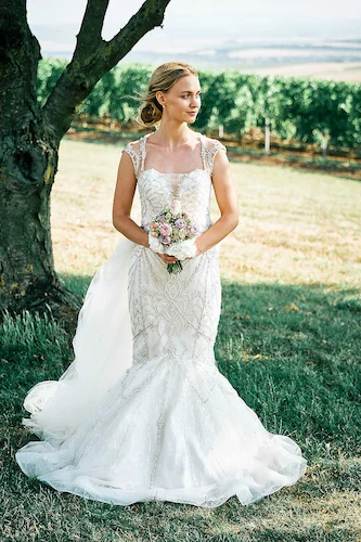 Onelovephoto Eine Braut in einem weißen, taillierten Hochzeitskleid hält einen Blumenstrauß, während sie im Freien auf einer Wiese in der Nähe eines Baumes steht, mit Weinbergen und einer malerischen Landschaft im Hintergrund. Hochzeitsfotografie in Mannheim und Heidelberg