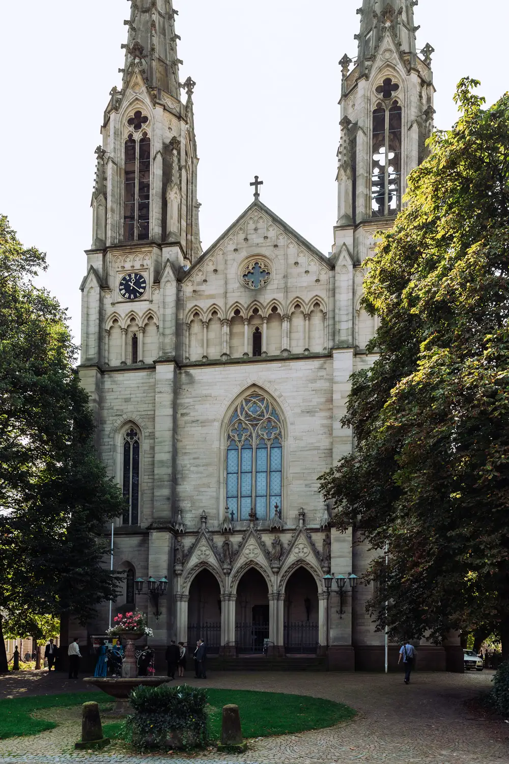 Eine hohe, historische Kirche mit zwei Türmen, großen Bogenfenstern und einer Uhr auf einem Turm steht in der Nähe von Brenners Spa in Baden-Baden Eine hohe, historische Kirche mit zwei Türmen, großen Bogenfenstern und einer Uhr auf einem Turm steht in der Nähe von Brenners Spa in Baden-Baden - Traumhochzeiten garantiert, umgeben von Bäumen und Spaziergängern. Onelovephoto - Hochzeitsfotografie in Mannheim und Heidelberg
