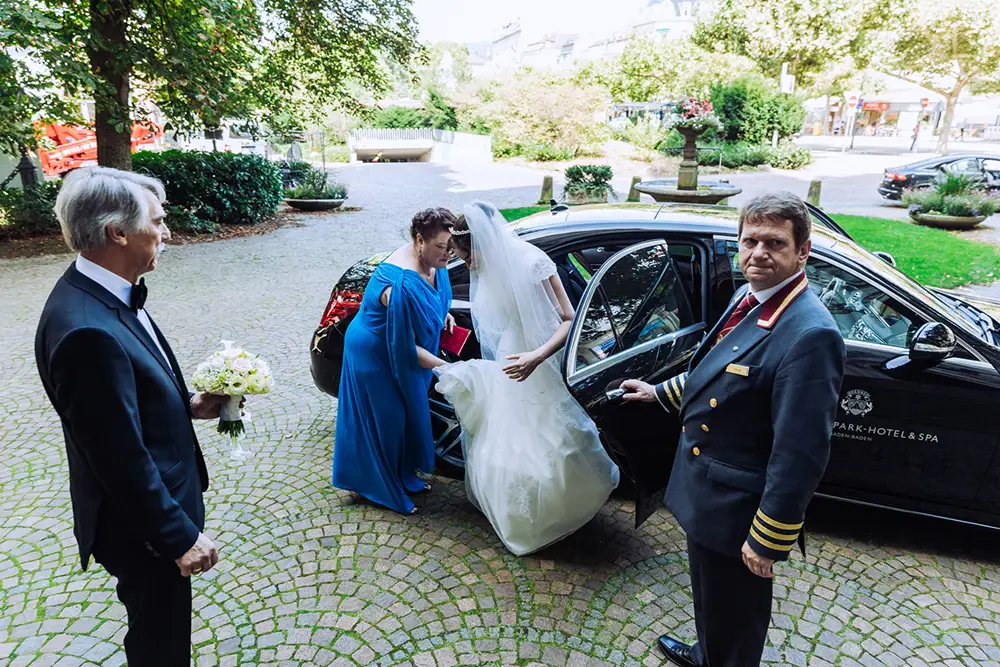 Eine Braut steigt aus einem schwarzen Auto, assistiert von einer Frau in Blau und einem Hotelportier. Ein älterer Mann im Smoking hält einen Blumenstrauß in der Hand und wartet in der Nähe auf einem Kopfsteinpflasterweg mit Grünpflanzen im Hintergrund. Onelovephoto - Hochzeitsfotografie in Mannheim und Heidelberg