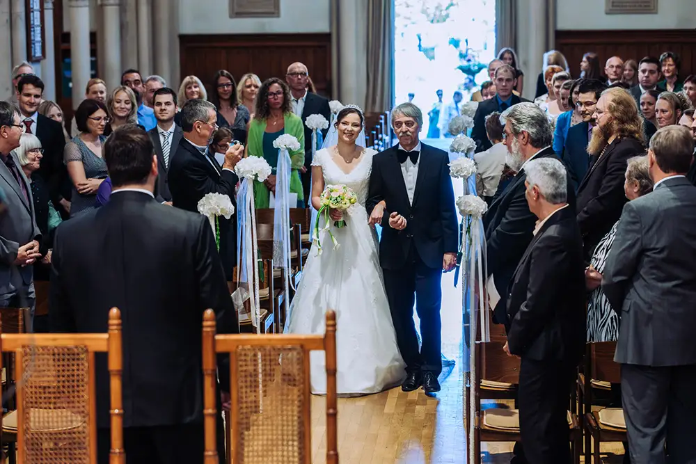 Eine Braut in einem weißen Kleid und mit einem Blumenstrauß in der Hand schreitet mit einem älteren Mann im Smoking den Gang hinunter, umgeben von stehenden Gästen bei einer Hochzeitszeremonie in einer Kirche. Onelovephoto - Hochzeitsfotografie in Mannheim und Heidelberg