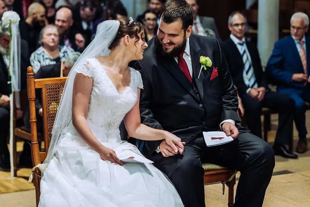 Braut und Bräutigam sitzen während der Hochzeitszeremonie eng beieinander, lächeln und halten sich an den Händen, umgeben von sitzenden Gästen in formellen Kleidern. Onelovephoto - Hochzeitsfotografie in Mannheim und Heidelberg