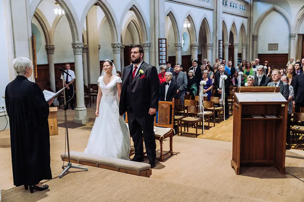 Braut und Bräutigam stehen während einer Hochzeitszeremonie in einer Kirche vor dem Altar, während die Gäste im Hintergrund sitzen und zusehen. Onelovephoto - Hochzeitsfotografie in Mannheim und Heidelberg