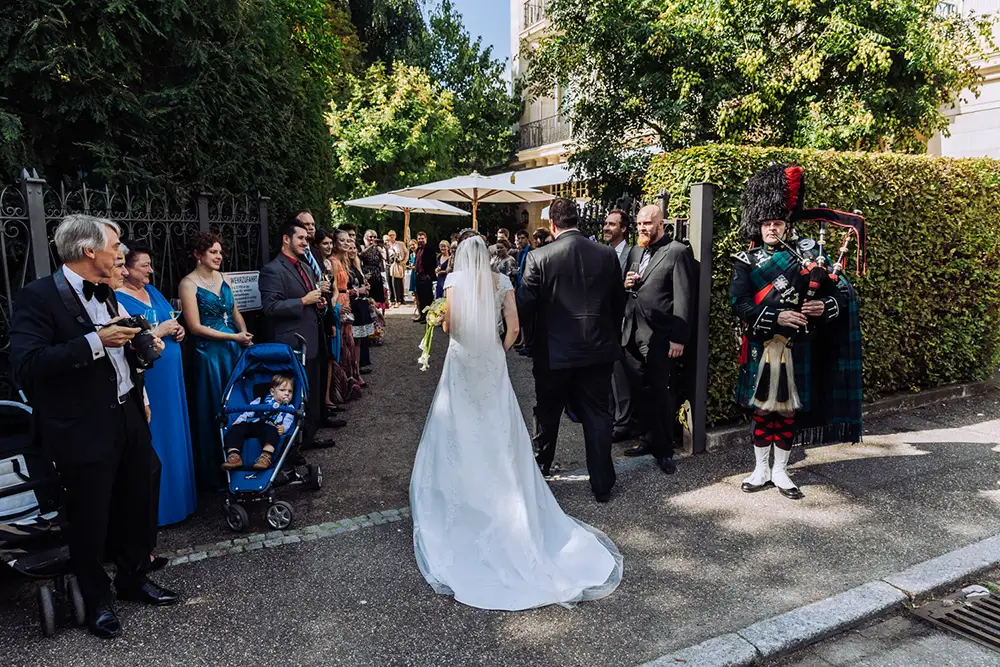 Eine Braut in einem weißen Kleid geht mit einem Mann auf die Gäste einer Hochzeitsfeier im Freien zu. Die Gäste, einige mit Kindern und Kinderwagen, stehen auf beiden Seiten, und ein Dudelsackspieler in traditioneller Kleidung spielt in der Nähe. Onelovephoto - Hochzeitsfotografie in Mannheim und Heidelberg
