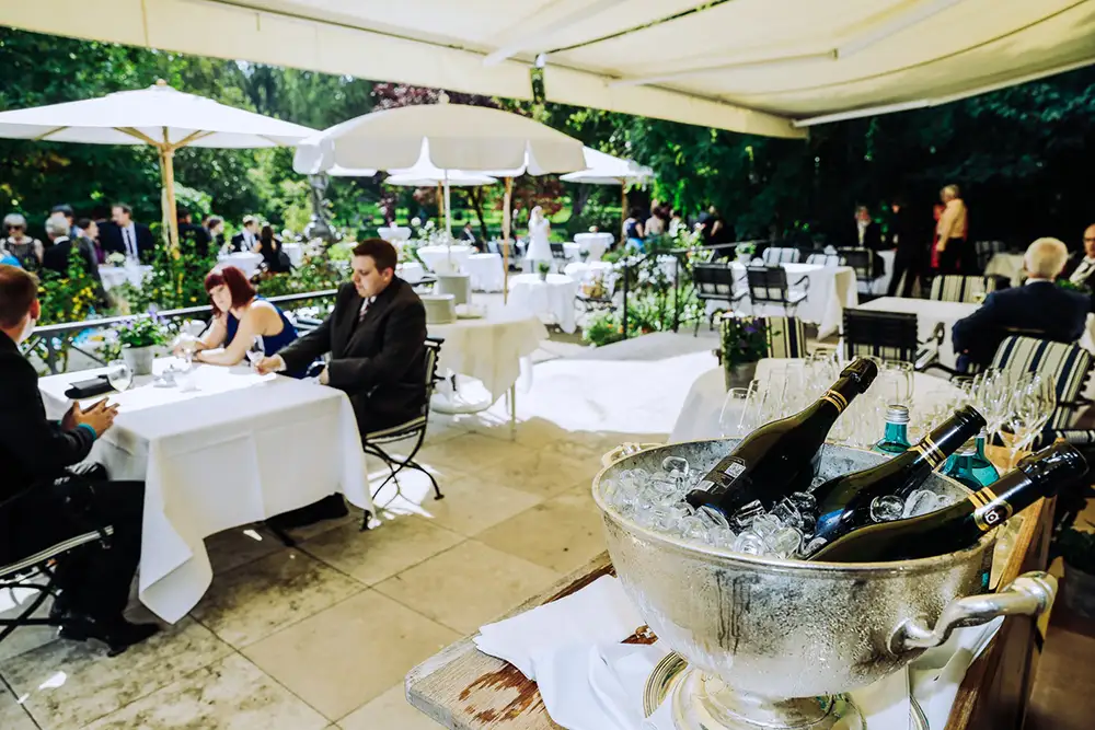 Bei einer Veranstaltung im Freien sitzen Menschen an weiß eingekleideten Tischen unter großen Sonnenschirmen. Im Vordergrund stehen Wein- und Sektflaschen in einem mit Eis gefüllten Eimer. Der Bereich ist von Grünflächen und Bäumen umgeben. Onelovephoto - Hochzeitsfotografie in Mannheim und Heidelberg