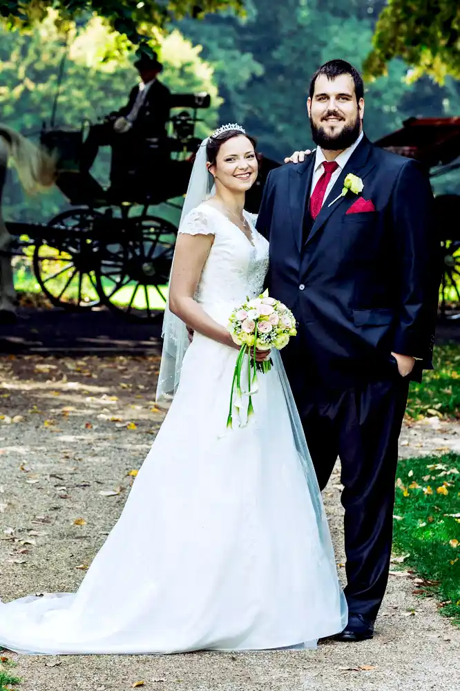 Eine Braut in einem weißen Kleid und ein Bräutigam in einem schwarzen Anzug mit roter Krawatte posieren lächelnd im Freien, während im unscharfen Hintergrund eine Pferdekutsche zu sehen ist. Eine Braut in einem weißen Kleid und ein Bräutigam in einem schwarzen Anzug mit roter Krawatte posieren lächelnd im Freien, während im unscharfen Hintergrund eine Pferdekutsche zu sehen ist.