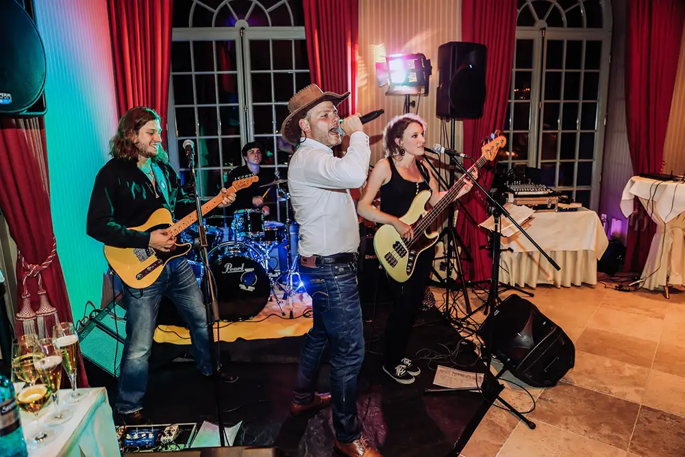 Eine vierköpfige Band tritt in einem Innenraum auf; der Sänger mit Cowboyhut singt energisch, während der Bassist, der Gitarrist und der Schlagzeuger spielen. Im Hintergrund sind rote Vorhänge und große Fenster zu sehen, und die Musikanlage umgibt sie. Onelovephoto - Hochzeitsfotografie in Mannheim und Heidelberg