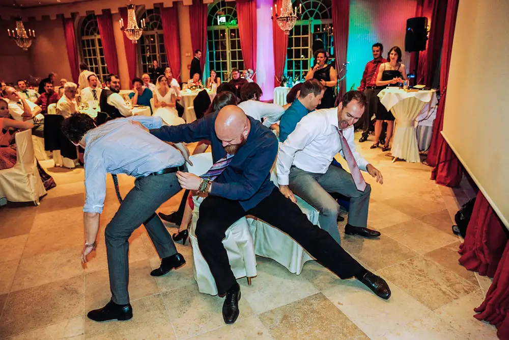 Eine lebhafte Gruppe Erwachsener spielt bei einer feierlichen Veranstaltung Reise nach Jerusalem, lacht und kämpft um die Plätze, während das Publikum, umgeben von elegantem Dekor und roten Vorhängen, zusieht. Onelovephoto - Hochzeitsfotografie in Mannheim und Heidelberg