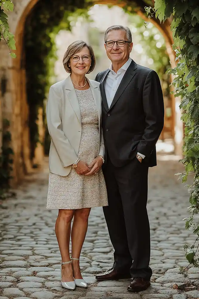 Ein älteres Paar steht dicht beieinander auf einem Kopfsteinpflasterweg, umgeben von Grünpflanzen und Steinbögen. Die Frau trägt ein helles Kleid und einen Blazer, während der Mann einen dunklen Anzug anhat. Beide lächeln in die Kamera. Onelovephoto - Hochzeitsfotografie in Mannheim und Heidelberg