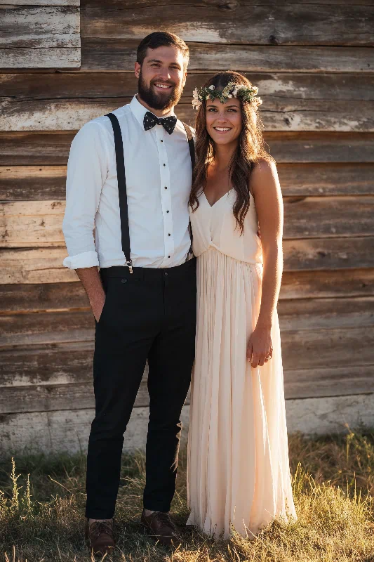 Ein lächelndes Paar steht vor einer rustikalen Holzwand. Der Mann trägt ein weißes Hemd, eine schwarze Hose, Hosenträger und eine Fliege. Die Frau trägt ein leichtes, fließendes Kleid und eine Blumenkrone. Das Sonnenlicht wirft einen warmen Schein auf die beiden.