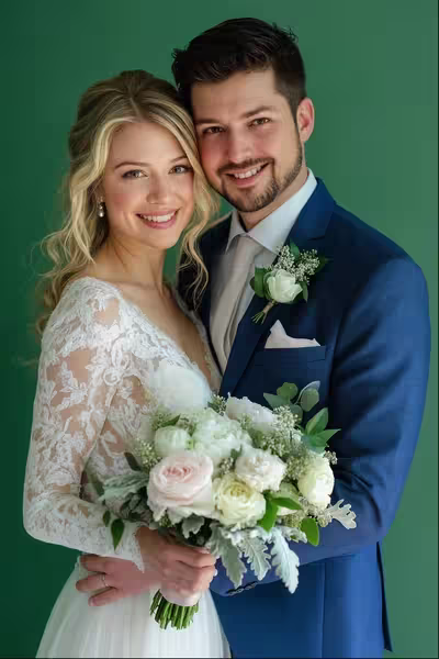 Eine lächelnde Braut im Spitzenhochzeitskleid hält einen Strauß weißer und rosafarbener Blumen, während sie neben einem Bräutigam im blauen Anzug mit Ansteckblume steht. Beide posieren vor einem grünen Hintergrund.