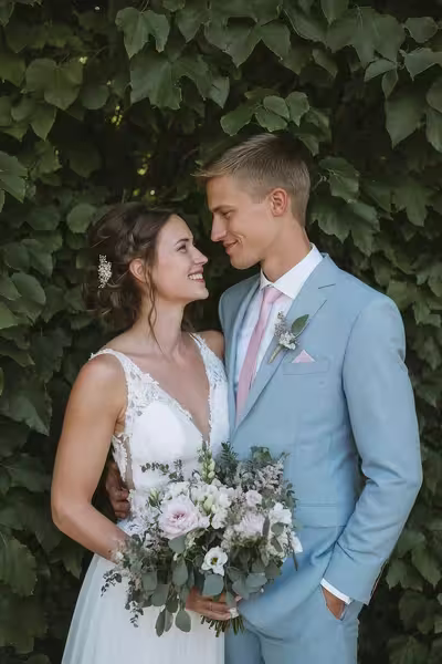 Eine Braut in einem weißen Kleid mit einem Blumenstrauß in der Hand und ein Bräutigam in einem hellblauen Anzug mit rosafarbener Krawatte stehen dicht beieinander und lächeln sich an, während im Hintergrund grünes Blattwerk zu sehen ist.