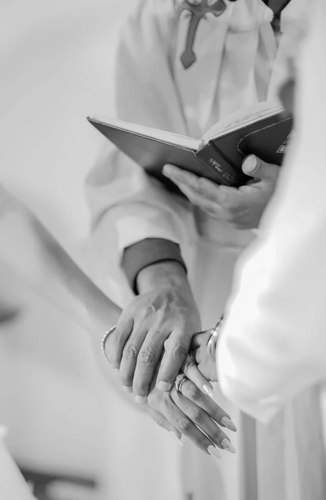 Ein Schwarz-Weiß-Foto mit drei übereinandergelegten Händen, während ein Priester ein aufgeschlagenes Buch in der Hand hält, deutet auf eine Hochzeit oder eine Segnungszeremonie hin. Die Szene wirkt feierlich und intim.