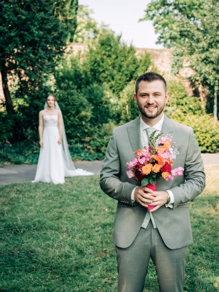 Der lächelnde Bräutigam Julius in einem hellgrauen Anzug hält einen bunten Blumenstrauß in der Hand. Janina, die Braut in einem weißen Kleid, steht verschwommen im Hintergrund, draußen, umgeben von viel Grün.