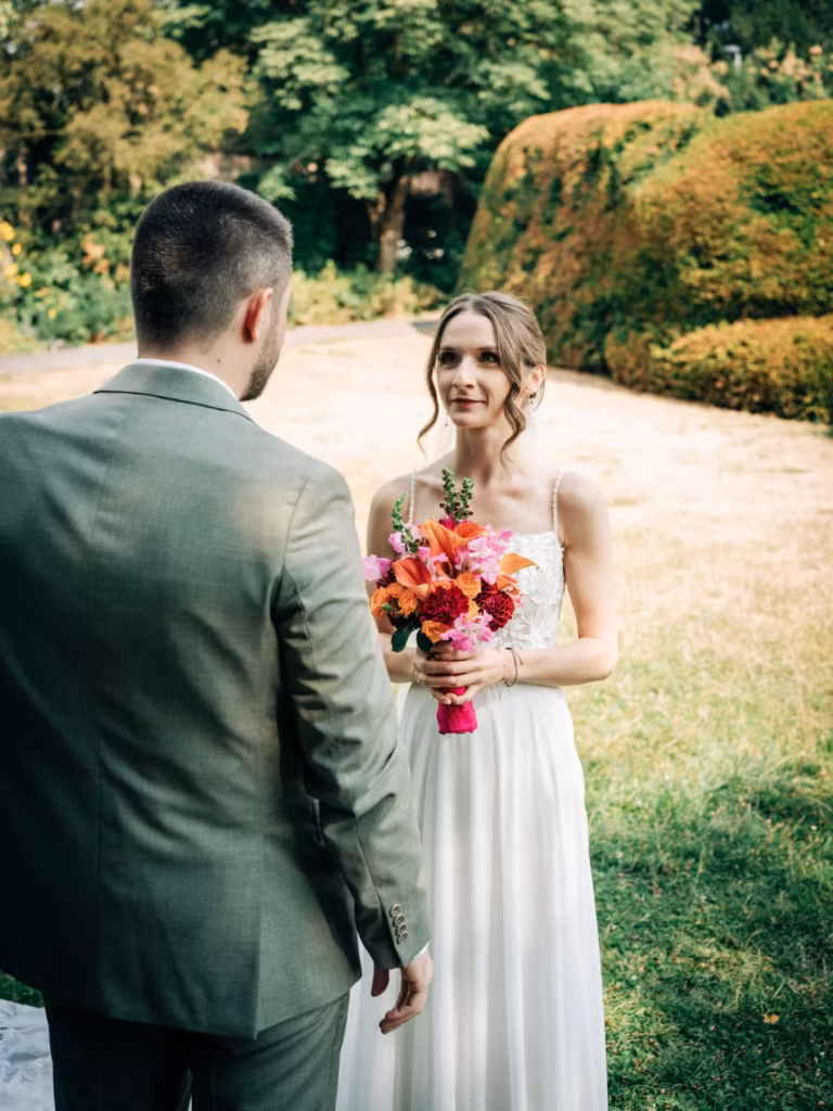 Eine Braut in einem weißen Kleid hält einen farbenfrohen Blumenstrauß in der Hand und lächelt den Bräutigam in einem grünen Anzug während einer Hochzeitszeremonie im Freien an, umgeben von viel Grün.