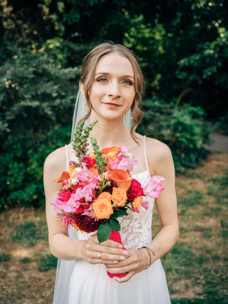 Eine Braut in einem weißen Kleid und mit Schleier steht im Freien und hält einen farbenfrohen Strauß aus rosa, orangefarbenen und roten Blumen in den Händen. Grünes Laub bildet den Hintergrund, perfekt eingefangen von einem Hochzeitsfotografen aus Weinheim.