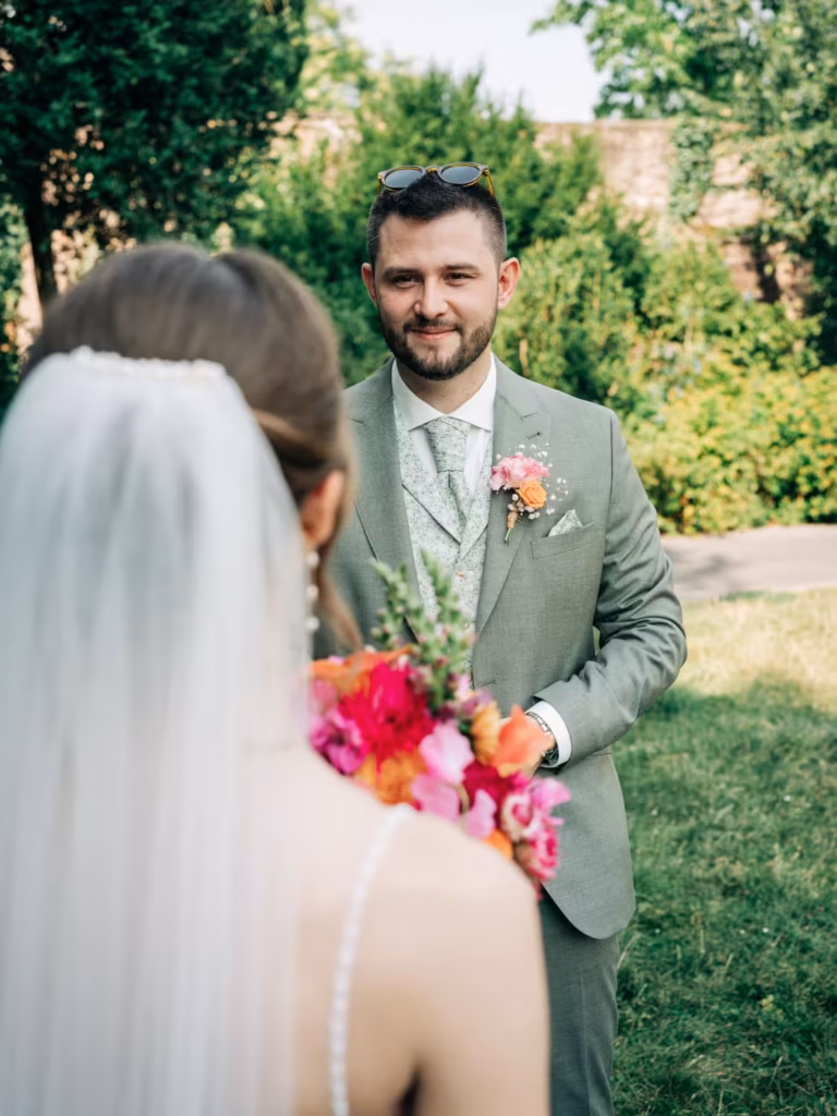 Ein Bräutigam in einem hellgrünen Anzug lächelt eine Braut in einem weißen Kleid und mit Schleier an, die einen bunten Blumenstrauß in der Hand hält; im Hintergrund ist eine grüne Landschaft zu sehen.