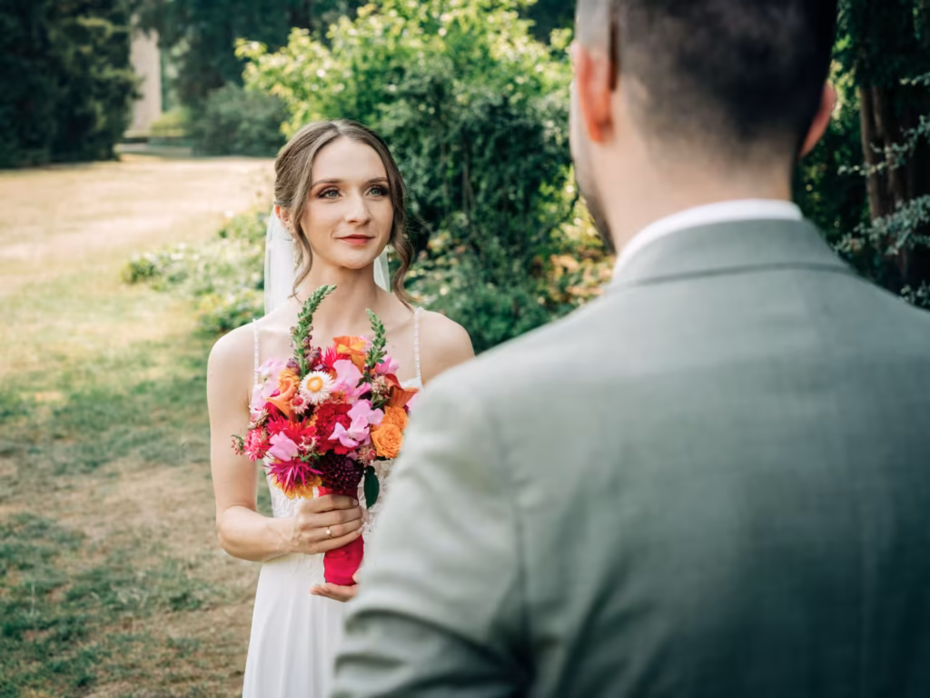 Eine Braut in einem weißen Kleid hält einen bunten Blumenstrauß in der Hand und lächelt einen Bräutigam in einem hellen Anzug im Freien an, umgeben von viel Grün – ein perfekter Moment, eingefangen von einem Hochzeitsfotografen aus Weinheim.