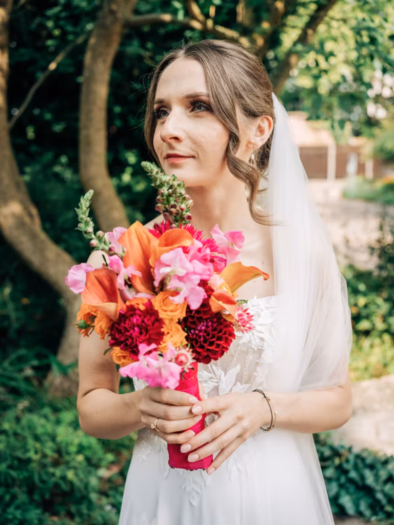 Eine Braut in einem weißen Kleid und mit Schleier hält einen bunten Strauß aus roten, rosa und orangefarbenen Blumen in der Hand und steht draußen vor dem Hintergrund von Bäumen und Grün – ein perfekter Moment, eingefangen von einem Hochzeitsfotografen aus Weinheim.