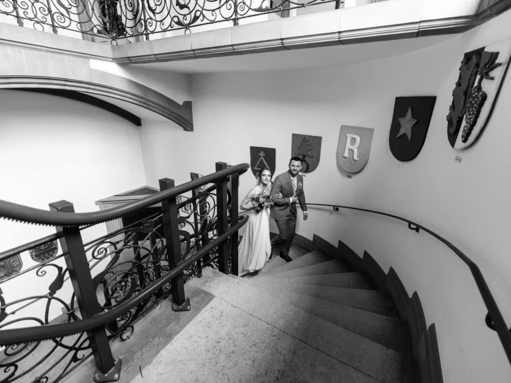 Eine Frau im Kleid und ein Mann im Anzug steigen in einem historischen Gebäude eine geschwungene Treppe hinauf, die mit kunstvollen Geländern und an den Wänden angebrachten Wappenschildern geschmückt ist – ein Bild, das die zeitlose Eleganz, für die der Hochzeitsfotograf Weinheim bekannt ist, perfekt einfängt.