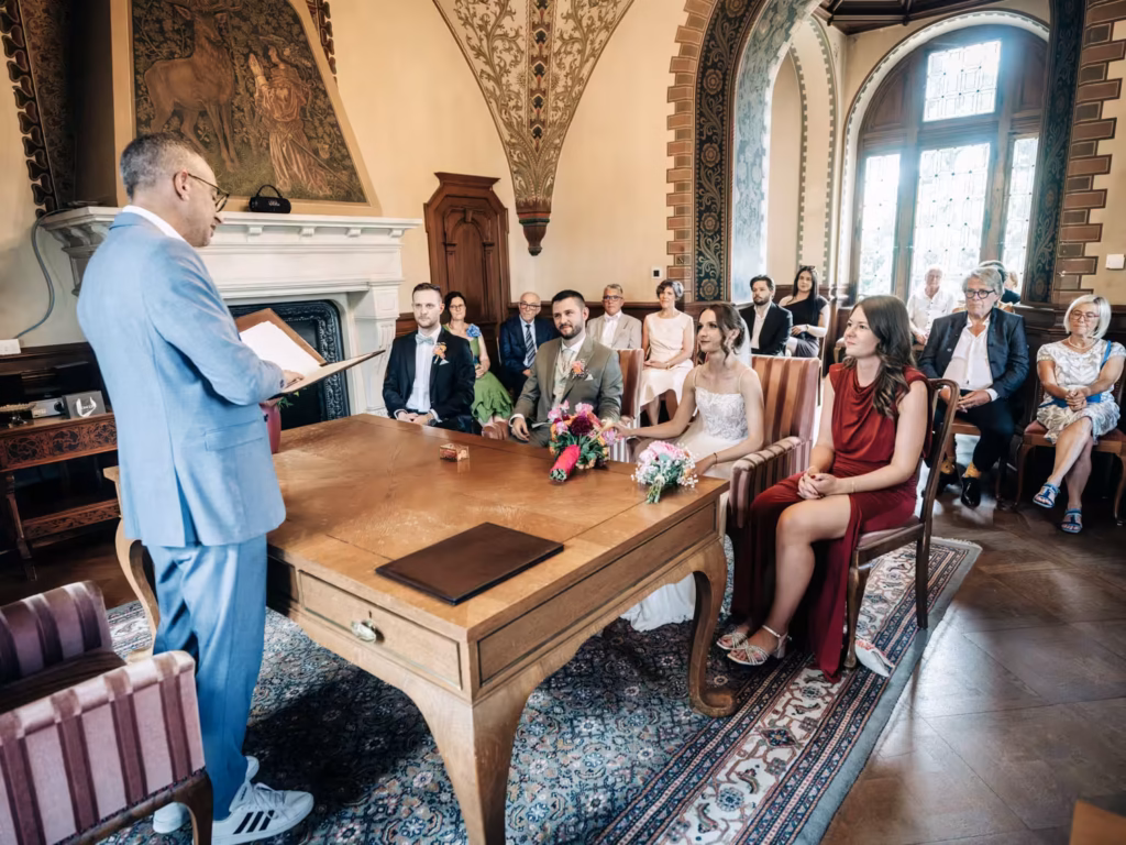 Ein Paar sitzt an einem Holztisch in einem prunkvoll geschmückten Raum und blickt auf einen Trauzeugen. Die Gäste verfolgen die Zeremonie, bei der es sich offenbar um eine Hochzeit oder eine standesamtliche Trauung handelt, vor dem Hintergrund von Blumen und eleganter Dekoration – wunderschön festgehalten von einem Hochzeitsfotografen aus Weinheim.