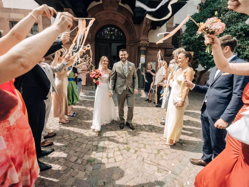 Ein Brautpaar geht Hand in Hand nach draußen, lächelt und ist umgeben von Gästen, die zur Feier des Tages mit Bändern winken. Die Sonne scheint, und alle sehen fröhlich aus – ein perfekter Moment, wunderschön festgehalten von einem Hochzeitsfotografen aus Weinheim.