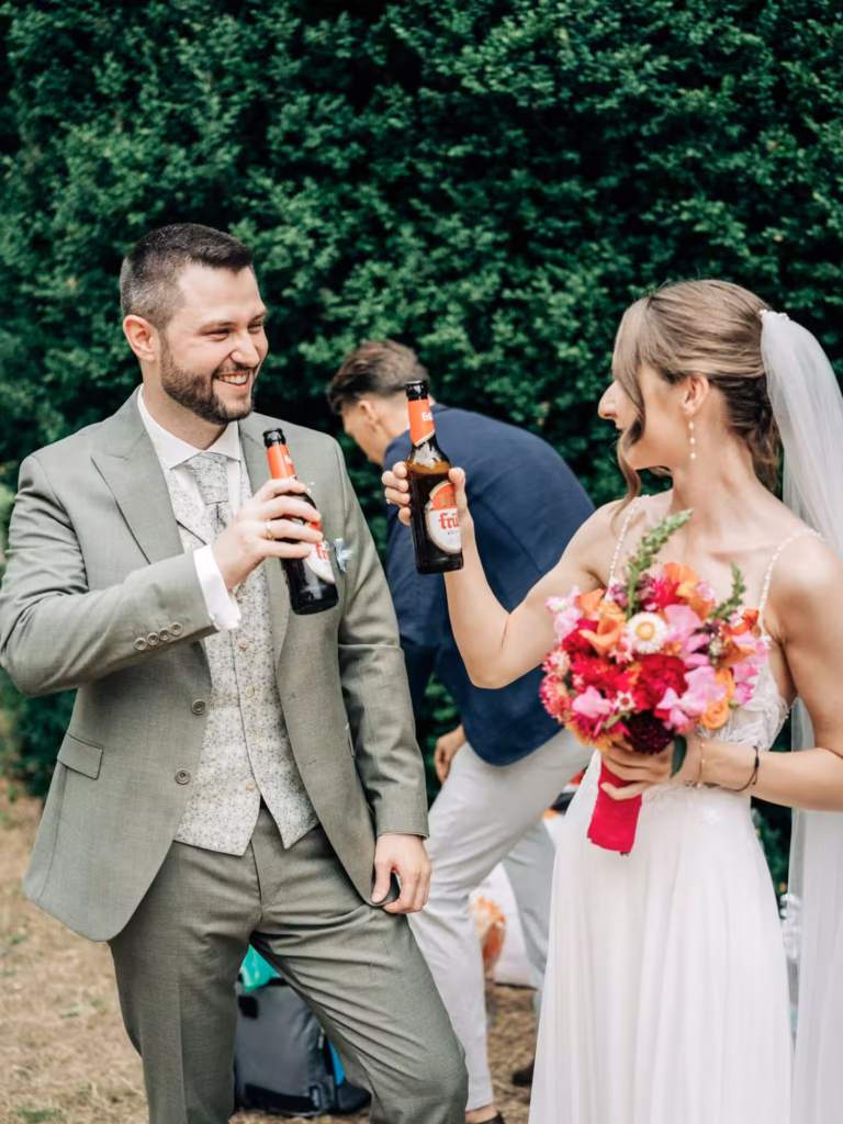 Ein Brautpaar lächelt und stößt im Freien mit Bierflaschen an. Die Braut hält einen Strauß rosa Blumen in der Hand. Im Hintergrund ist ein Mann in einer blauen Jacke zu sehen, der teilweise abgewandt steht. Grüne Büsche füllen den Hintergrund aus.