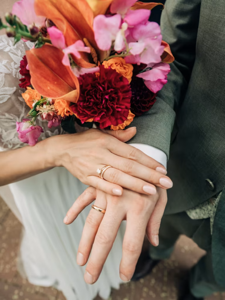 Zwei Personen in Hochzeitskleidung zeigen ihre Hände mit Eheringen, wobei eine Person einen farbenfrohen Strauß aus rosa, orangefarbenen und roten Blumen in der Hand hält.