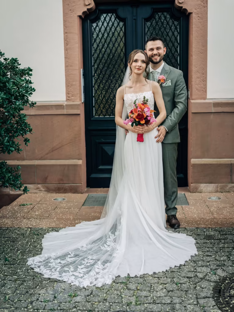 Eine Braut in einem weißen Kleid, die einen Blumenstrauß in der Hand hält, steht vor einem Bräutigam in einem grauen Anzug; gemeinsam posieren sie lächelnd vor einem Gebäude mit einer schwarzen Tür und etwas Grün im Hintergrund – perfekt eingefangen von einem Hochzeitsfotografen aus Weinheim.