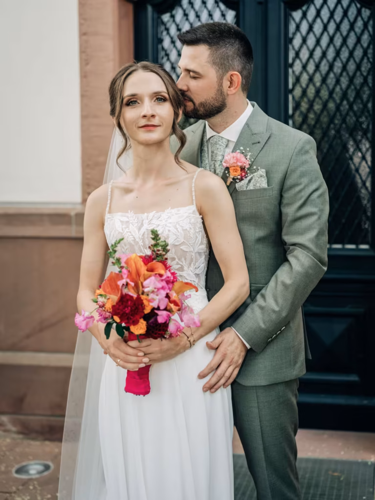 Eine Braut in einem weißen Kleid, die einen bunten Blumenstrauß in der Hand hält, steht vor einem Bräutigam im grünen Anzug, der sie sanft umarmt und ihr einen Kuss auf die Stirn gibt – ein zeitloser Moment, wunderschön festgehalten vom Hochzeitsfotografen Weinheim.