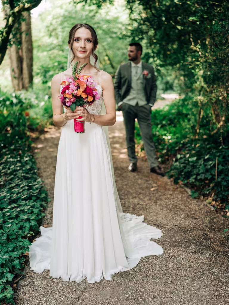 Eine Braut in einem weißen Kleid, die einen bunten Blumenstrauß in der Hand hält, steht auf einem Gartenweg, während hinter ihr ein Mann im Anzug steht, umgeben von viel Grün.
