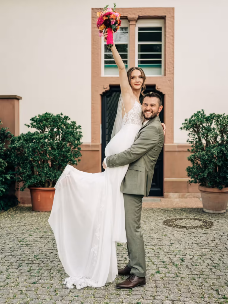 Ein Bräutigam im grünen Anzug hebt seine Braut, die ein weißes Kleid trägt und einen Blumenstrauß über den Kopf hält, lächelnd vor einem Gebäude mit Topfpflanzen in die Luft – ein Bild, das die Freude und Romantik perfekt einfängt, wie es durch das Objektiv eines Hochzeitsfotografen aus Weinheim zu sehen ist.