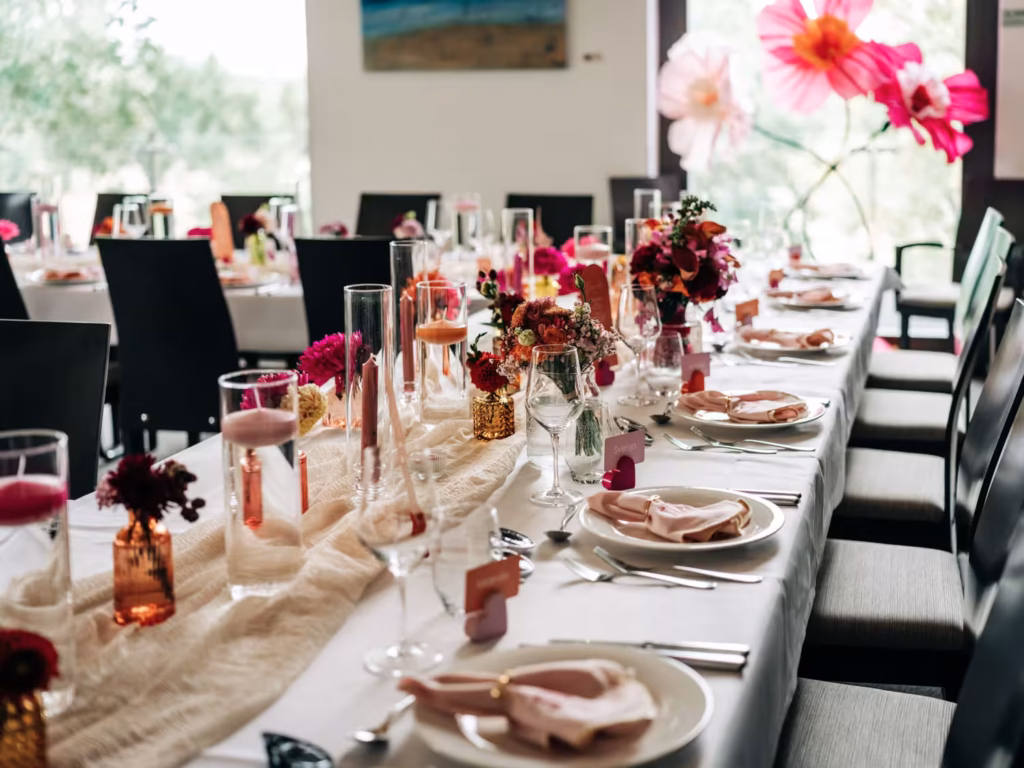 Ein elegantes Tischarrangement für eine Veranstaltung mit Dekoration in Rosa und Gold, Blumengestecken, Kerzen, Gläsern, Tellern und Servietten, umgeben von schwarzen Stühlen in einem hellen Raum mit großen Fenstern und üppigen Blumendekorationen – eine perfekte Kulisse, die ein Hochzeitsfotograf aus Weinheim festhalten kann.