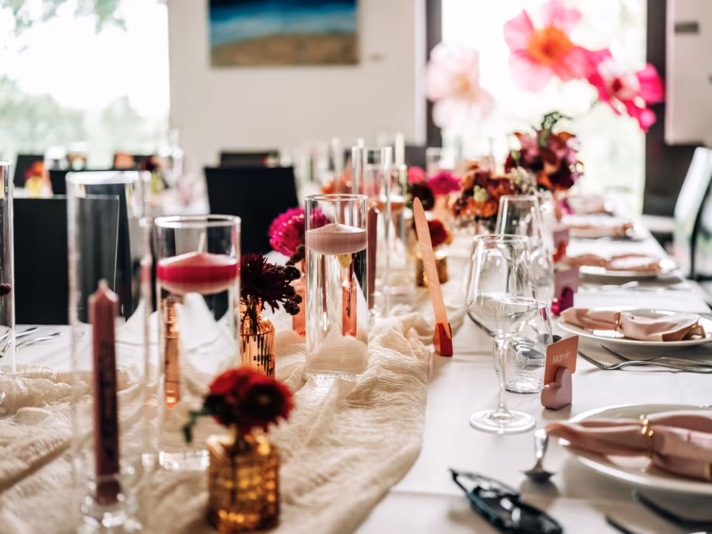 Ein langer Esstisch ist elegant mit rosa Kerzen, Weingläsern, gefalteten Servietten und roten Blumengestecken gedeckt und schafft so eine festliche und romantische Atmosphäre – perfekt, um unvergessliche Momente mit einem Hochzeitsfotografen aus Weinheim festzuhalten.