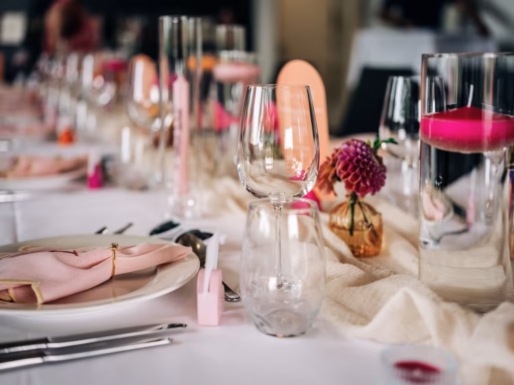 Eine Nahaufnahme eines elegant gedeckten Tisches mit rosa Servietten, Gläsern, Besteck und rosa Blumen in kleinen goldenen Vasen, die auf einer weißen Tischdecke arrangiert sind.