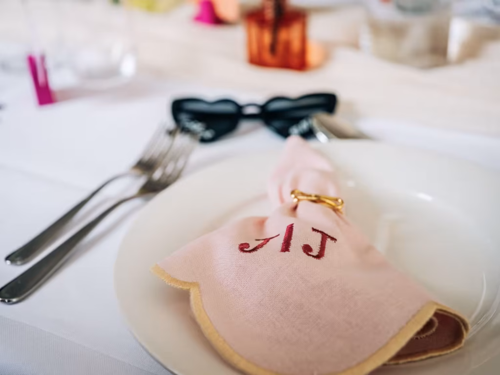 Eine Nahaufnahme einer Tischdekoration mit einer rosa Serviette, auf der „JJ“ gestickt ist; sie ist in einen goldenen Ring gewickelt und liegt auf einem weißen Teller neben Besteck und einer schwarzen Sonnenbrille.