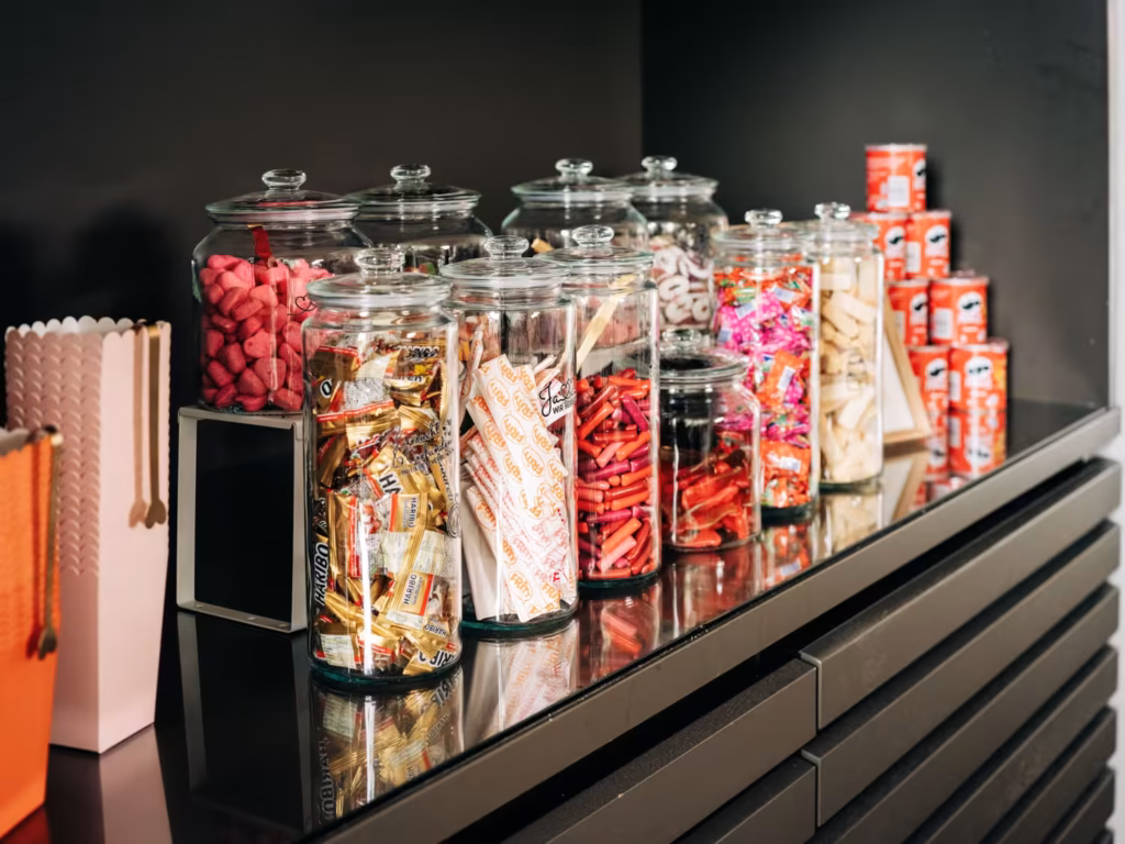 Auf einer Arbeitsplatte stehen Glasgefäße, die mit verschiedenen Süßigkeiten und Snacks gefüllt sind. In der Ecke sind kleine Pringles-Dosen gestapelt. Außerdem ist auf der linken Seite des Bildes eine weiße Popcorn-Schachtel zu sehen.