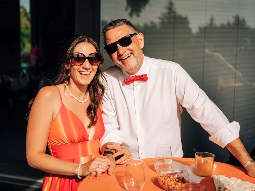 Ein lächelnder Mann und eine lächelnde Frau mit Sonnenbrillen sitzen an einem orangefarbenen Tisch mit Snacks und Getränken, sind halbformell gekleidet und genießen eine sonnige Veranstaltung im Freien.