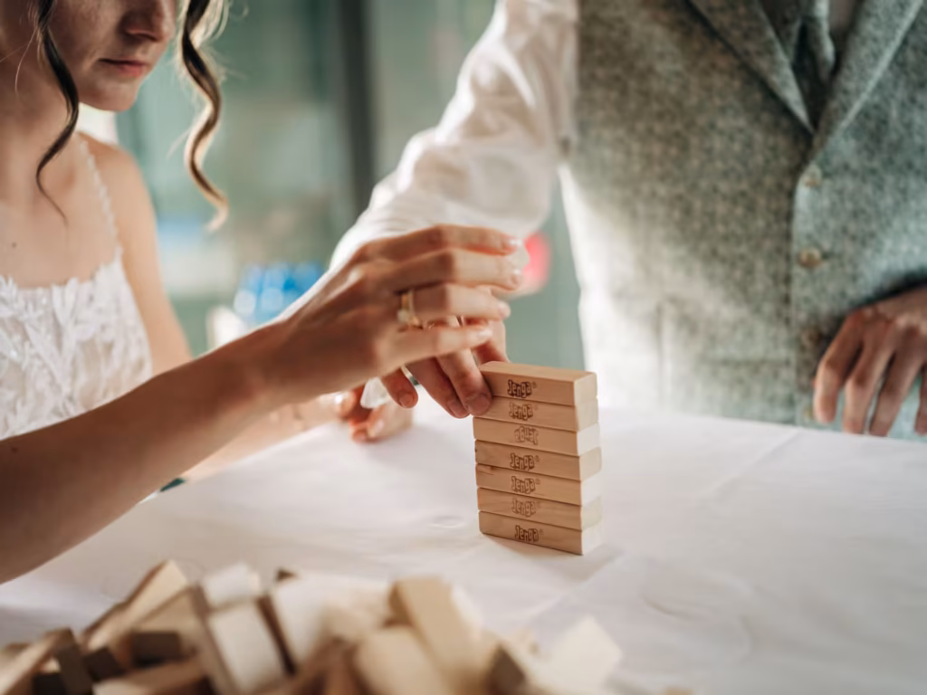 Ein Brautpaar in Hochzeitskleidung spielt gemeinsam an einem Tisch eine Partie Jenga und zieht vorsichtig einen Block aus dem aufgestapelten Turm heraus.