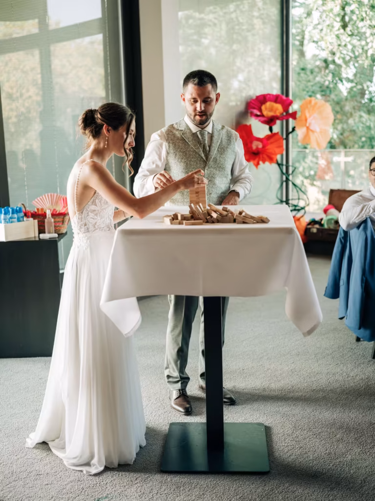Ein Brautpaar in Hochzeitskleidung spielt drinnen an einem hohen Tisch eine Partie Jenga, im Hintergrund sind große, bunte Papierblumen zu sehen.
