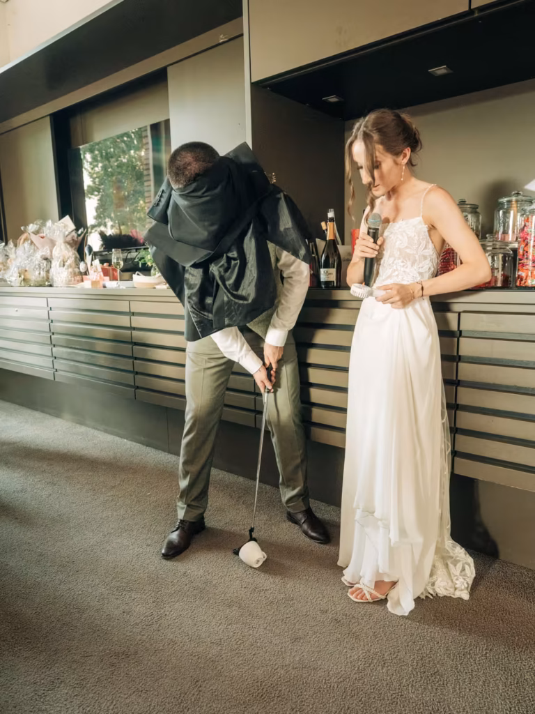 Eine Person in einem weißen Kleid hält ein Mikrofon in der Hand, während eine andere Person, deren Kopf mit schwarzem Stoff bedeckt ist, versucht, mit einem Golfschläger auf einen kleinen Gegenstand auf dem Boden zu schlagen.