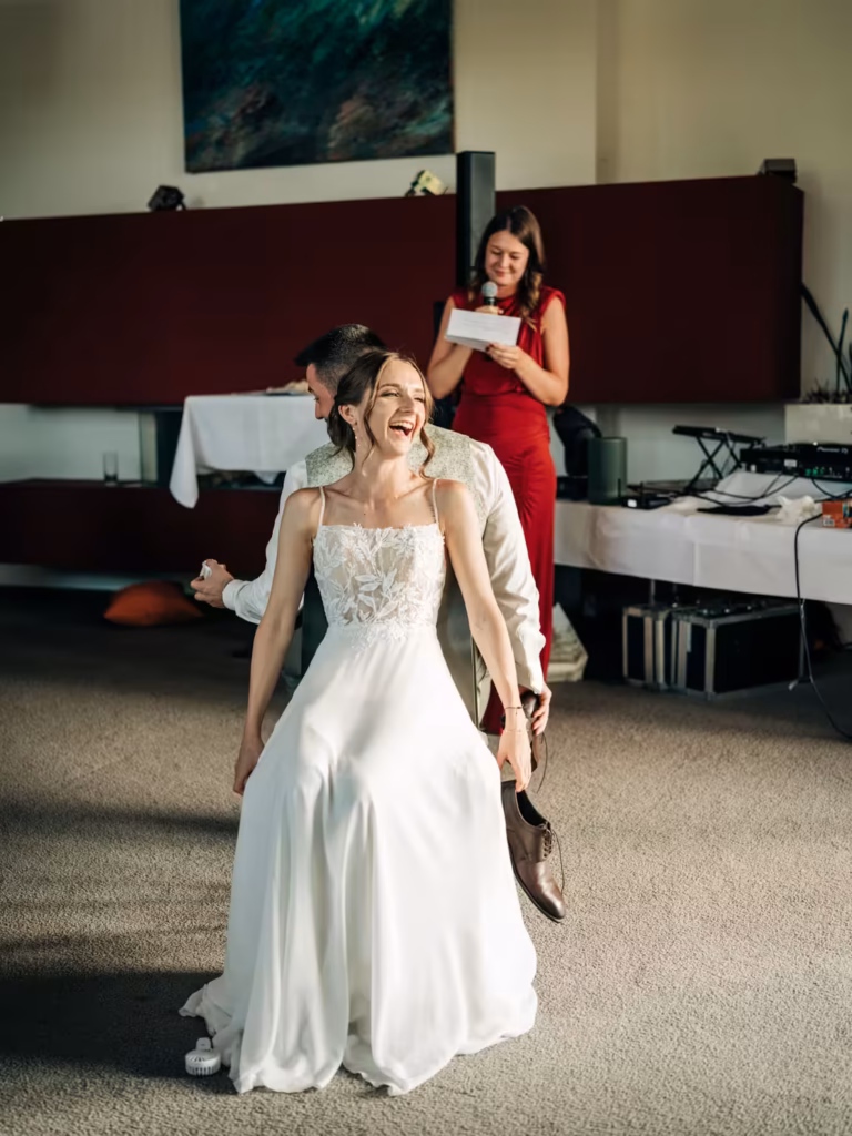 Eine Braut in einem weißen Kleid lacht, während sie auf dem Schoß ihres Bräutigams sitzt; hinter ihnen steht eine Frau in einem roten Kleid und liest von einer Karte vor.