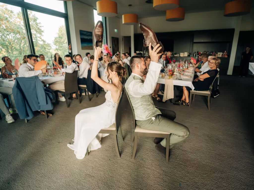 Bei einem Spiel während der Hochzeitsfeier sitzen Braut und Bräutigam Rücken an Rücken und halten sich gegenseitig die Schuhe hoch, umgeben von Gästen an festlich geschmückten Tischen in einem hellen, modernen Veranstaltungsraum.
