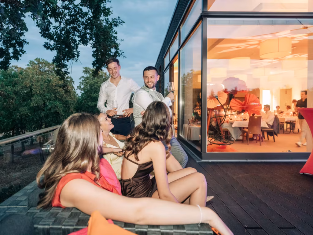 Vier Personen sitzen auf einem Sofa auf der Terrasse, unterhalten sich und lachen, während zwei Personen in Hemden daneben stehen. Durch große Fenster sieht man weitere Menschen, die in einem hell erleuchteten, modernen Gebäude zu Abend essen.