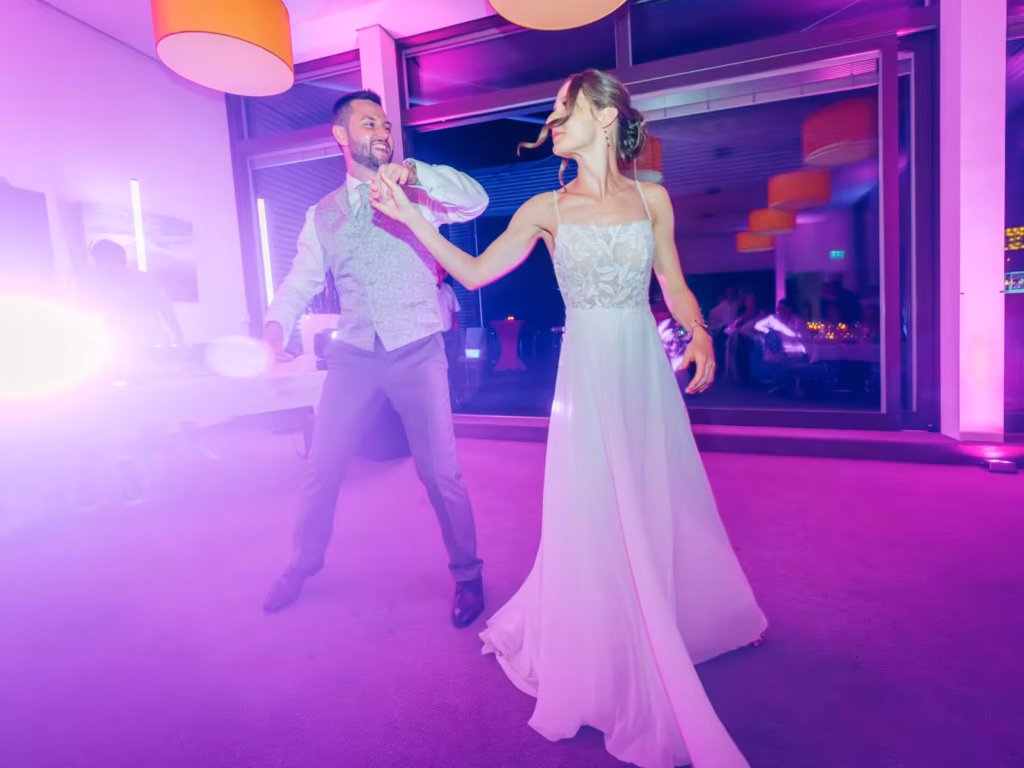 Eine Braut in einem weißen Brautkleid und ein Mann in Abendgarderobe tanzen fröhlich miteinander in einem hell erleuchteten, farbenfrohen Raum, der von violettem Licht und hängenden Lampen erhellt wird.
