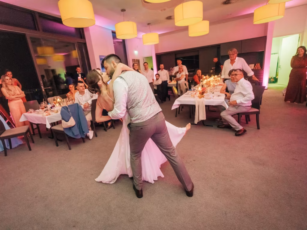 Ein Brautpaar tanzt romantisch in einem warm beleuchteten Raum, umgeben von sitzenden Gästen, die zuschauen und lächeln, mit festlich gedeckten Tischen und sanftem rosa Licht.