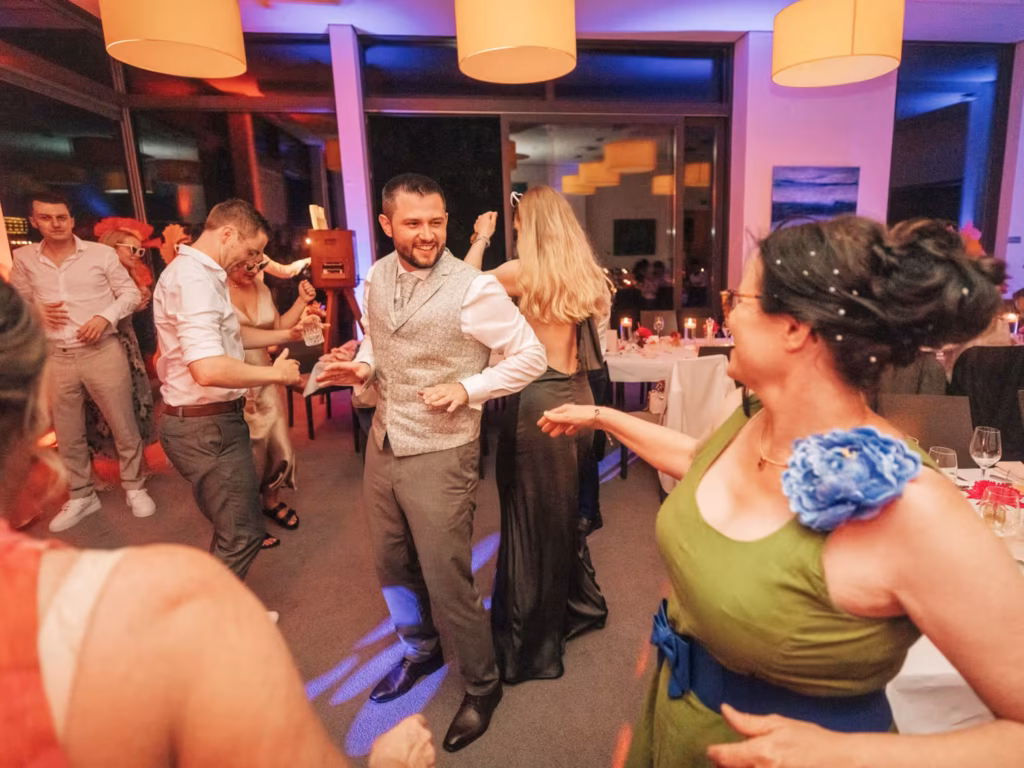 Auf einer lebhaften Party in einem Innenraum mit bunten Lichtern und runden Deckenlampen tanzen die Gäste und lächeln. Im Hintergrund stehen Tische mit Getränken und Dekorationen. Alle scheinen die Feier zu genießen.