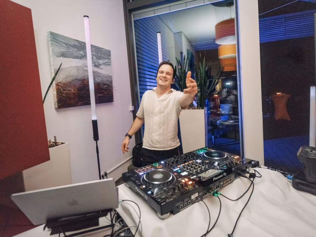 Ein lächelnder DJ in einem weißen Hemd steht hinter einem Pioneer-DJ-Setup und streckt eine Hand in Richtung Kamera aus. Im Hintergrund sind eine moderne Einrichtung und blaue Beleuchtung zu sehen.