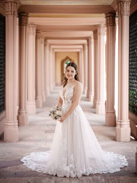 Eine Braut in einem weißen Brautkleid hält einen Blumenstrauß in der Hand und steht in einem eleganten Flur, der von hohen Säulen gesäumt ist und in sanftem Licht erstrahlt.