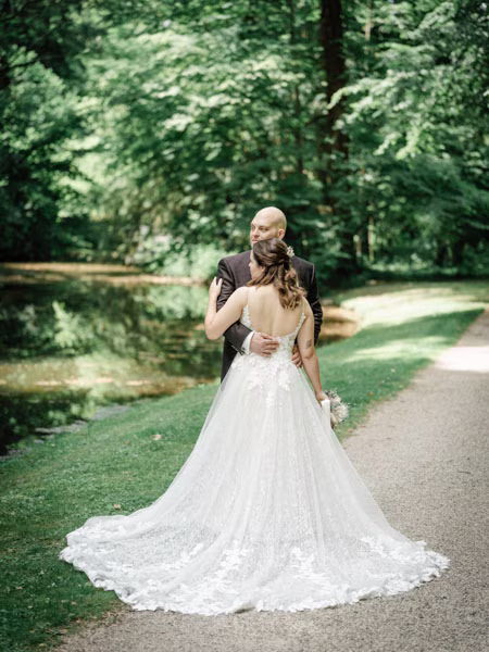 Eine Braut in einem weißen Brautkleid steht mit einem Mann im Anzug an einem Teich, umgeben von Grün. Sie stehen mit dem Rücken zur Kamera und umarmen sich auf einem sonnenbeschienenen Weg.